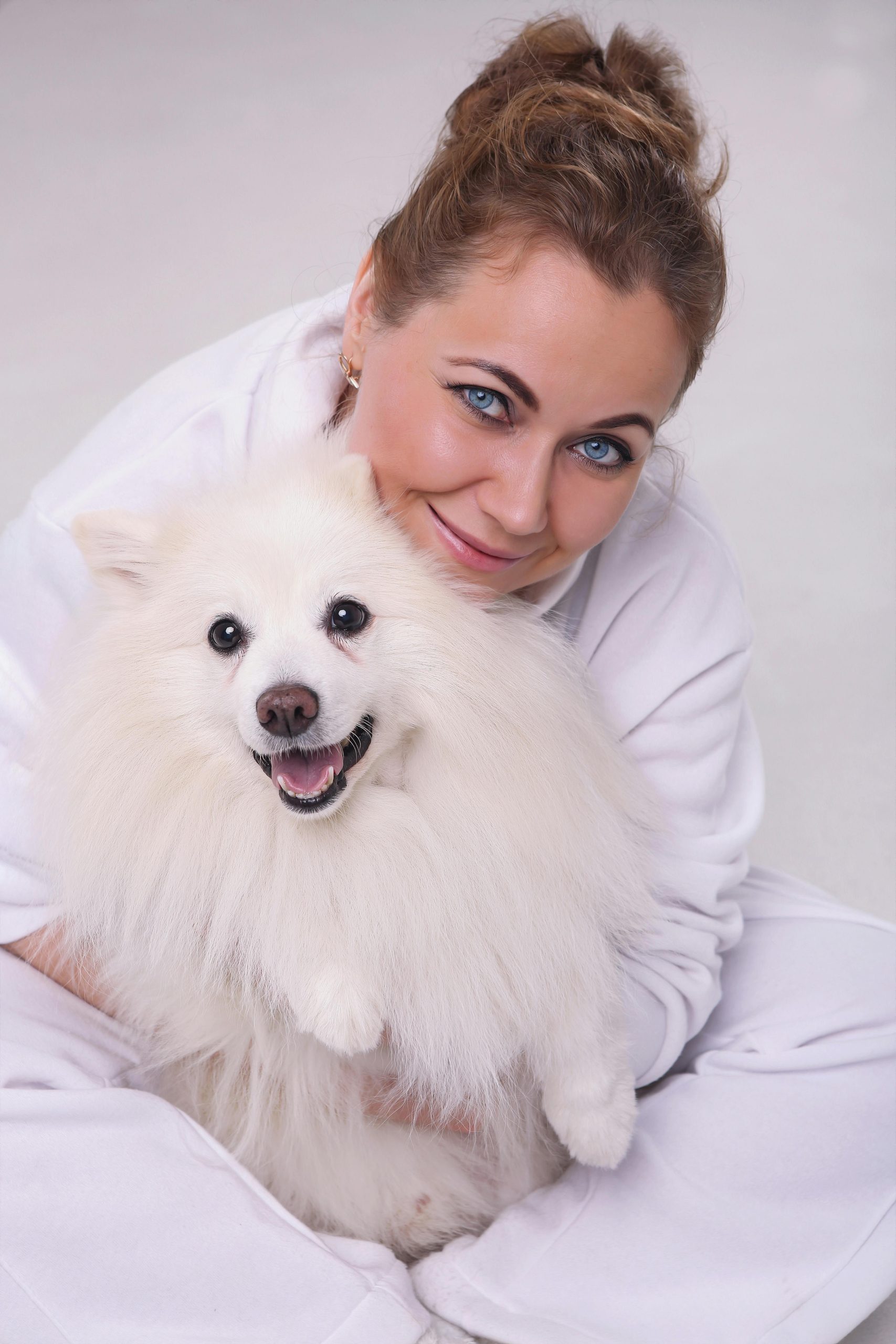 A woman in white happily embraces her smiling Pomeranian dog indoors.